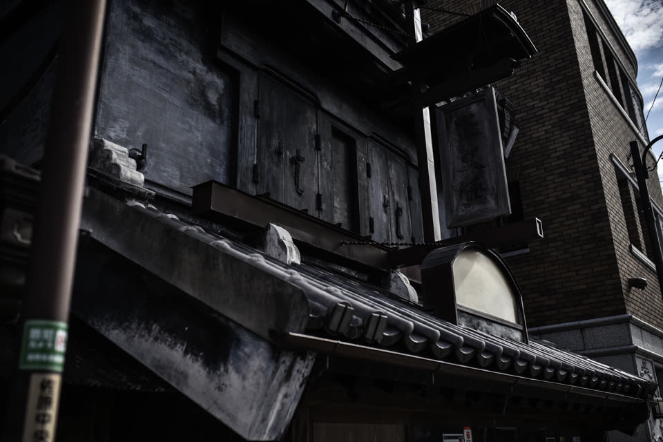見上げた黒く風化した木造の庇。右奥に煉瓦の建物と空