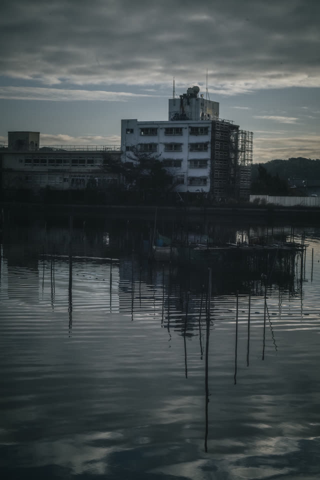 対岸の建物と、細波の立つ水面に割れて映る影。空は曇り