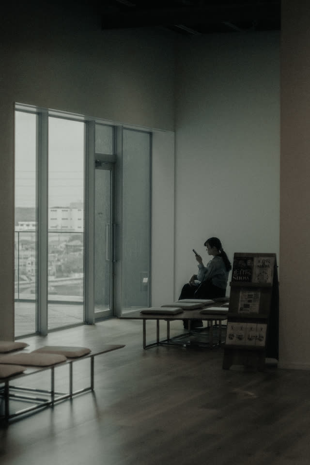 明るい室内の壁際のベンチに一人。奥に大きな窓と低い机